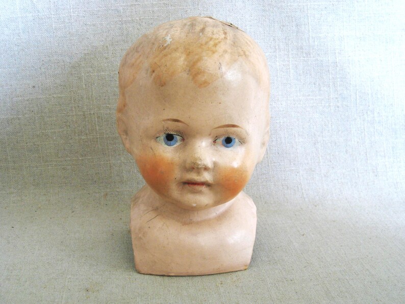 May include: Antique doll head with blue eyes and rosy cheeks. The doll head has blonde hair and a neutral skin tone. The doll head is a vintage item, likely porcelain or bisque.