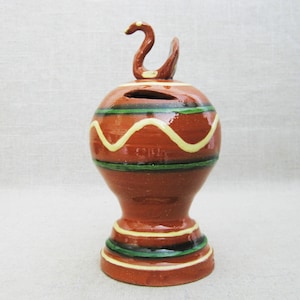 May include: A brown ceramic piggy bank with a green and yellow striped design. The piggy bank has a bird-shaped lid with a hole for coins.