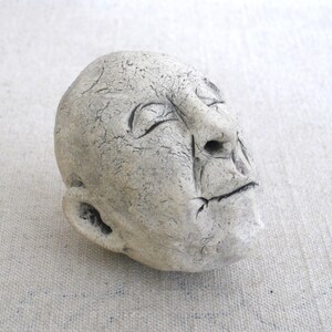 May include: A white ceramic sculpture of a face with closed eyes and a slightly open mouth. The sculpture has a textured surface and appears to be made of rough clay.