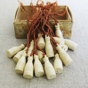 May include: A collection of small, white, bottle-shaped charms with brown strings attached. The charms are arranged in a box with a brown and white patterned interior.