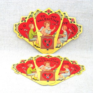 May include: Vintage Valentine's Day card with a pop-up design. The card features a cupid with wings, holding a book and pointing to a heart that says "To My Valentine". Two children are sitting at desks on either side of the cupid, with hearts that say "Love You" on their desks.
