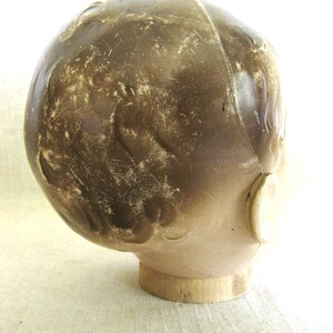 May include: A vintage doll head with a brown, aged appearance. The head is mounted on a wooden base. The doll head has a light skin tone and a dark brown, textured surface, with visible wear and tear.