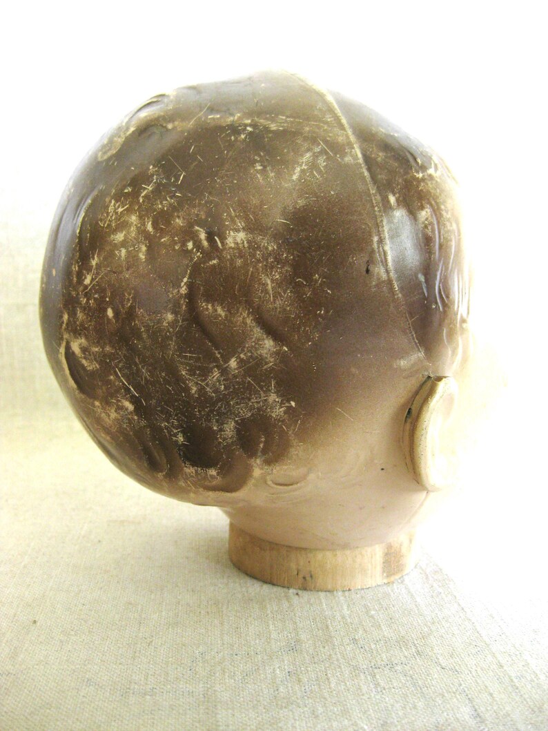 May include: A vintage doll head with a brown, aged appearance. The head is mounted on a wooden base. The doll head has a light skin tone and a dark brown, textured surface, with visible wear and tear.