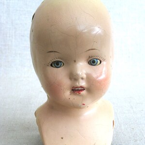 Puede incluir: Una cabeza de mu&ntilde;eca de porcelana vintage con ojos azules y una superficie agrietada. La cabeza de la mu&ntilde;eca no tiene cuerpo y tiene una boca roja pintada.