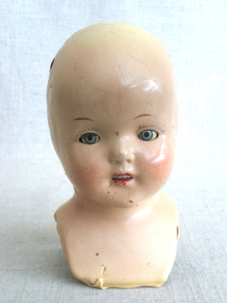 Puede incluir: Una cabeza de mu&ntilde;eca de porcelana vintage con ojos azules y una superficie agrietada. La cabeza de la mu&ntilde;eca no tiene cuerpo y tiene una boca roja pintada.