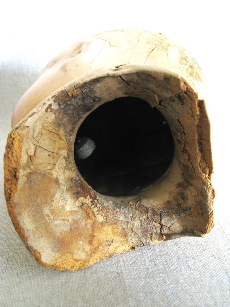 May include: A vintage, hollow ceramic head with a large, circular opening. The head is a light beige color with visible cracks and imperfections. The interior of the head is dark, and the edges show signs of age and wear.