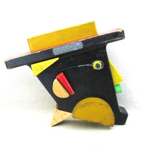 May include: Abstract wooden sculpture with geometric shapes in black, yellow, red, and green. The sculpture has a face-like design with a yellow nose and a red cylindrical element. The text "A1-377" is visible on the top.