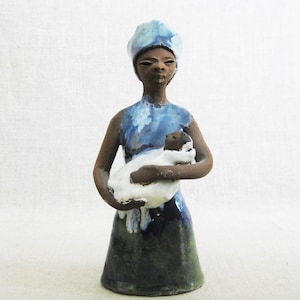 May include: A ceramic figurine of a woman wearing a blue and white head covering, holding a white ceramic lamb in her arms. The figurine is glazed in shades of blue and green.