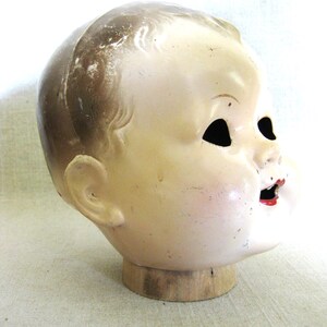 May include: Vintage doll head with a painted face, mounted on a wooden base. The doll head has a pale complexion, with black eye sockets and red lips. The hair is a light brown color. The head is mounted on a round wooden base.