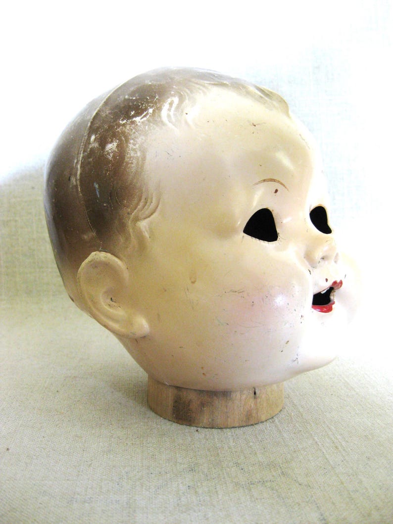 May include: Vintage doll head with a painted face, mounted on a wooden base. The doll head has a pale complexion, with black eye sockets and red lips. The hair is a light brown color. The head is mounted on a round wooden base.
