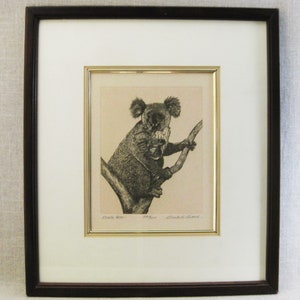 May include: Framed black and white print of a koala bear perched on a tree branch. The artwork is in a gold frame, set against a white mat, and enclosed in a dark brown wooden frame. The print is signed and includes the text "Koala bear".