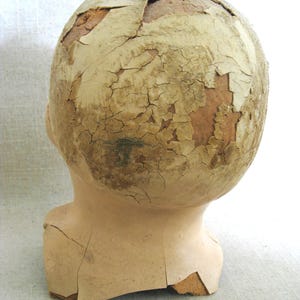May include: A vintage doll head with a cracked, peeling surface. The head is a light beige color, with the top of the head showing significant damage and exposed material. The neck is intact, and the overall appearance suggests age and wear.