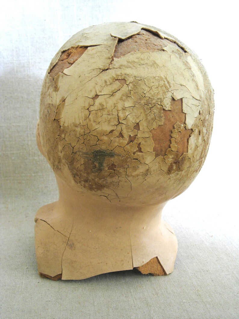 May include: A vintage doll head with a cracked, peeling surface. The head is a light beige color, with the top of the head showing significant damage and exposed material. The neck is intact, and the overall appearance suggests age and wear.