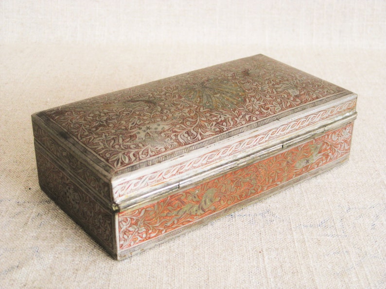 May include: A rectangular silver box with intricate floral designs and red accents. The box has a hinged lid and is decorated with a silver border.