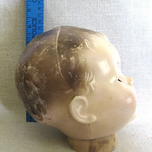 May include: Vintage doll head, side profile, with light skin tone and brown hair. The doll head is mounted on a wooden base. A blue ruler with the word "POLICE" is visible in the background.