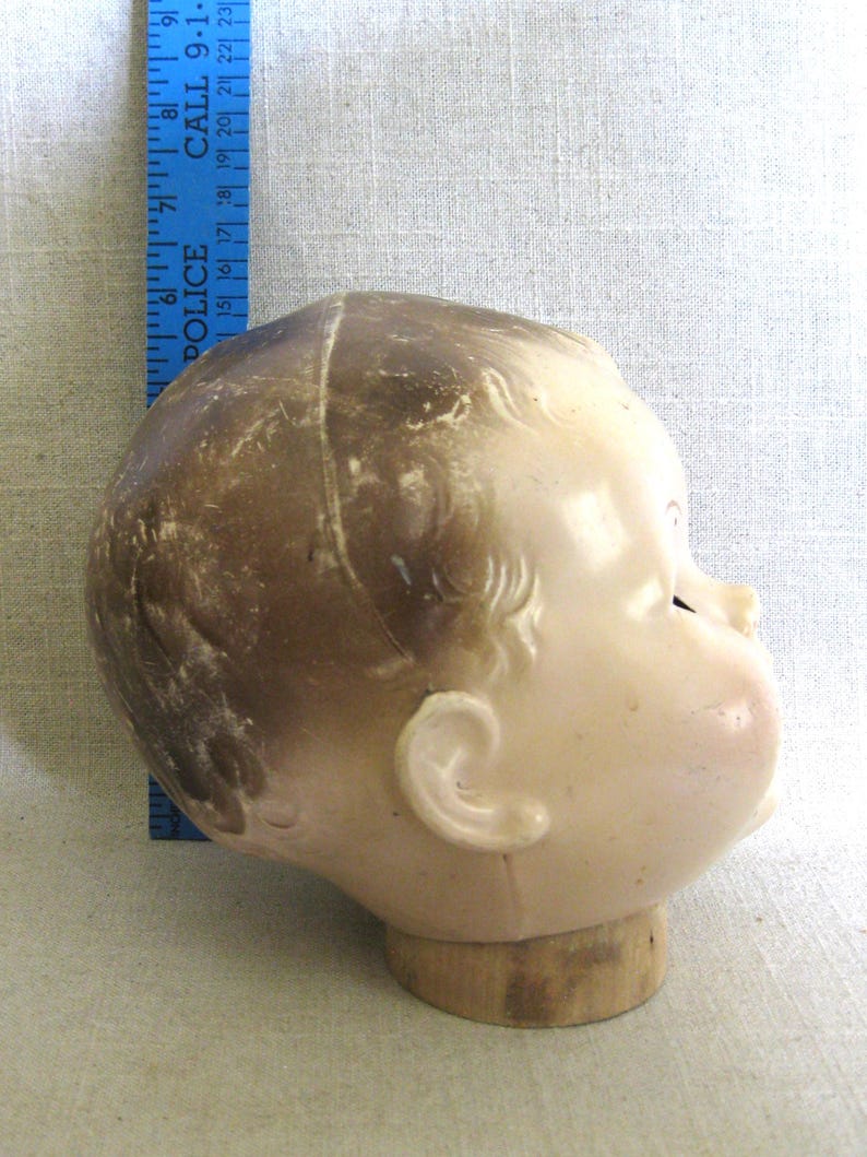 May include: Vintage doll head, side profile, with light skin tone and brown hair. The doll head is mounted on a wooden base. A blue ruler with the word "POLICE" is visible in the background.