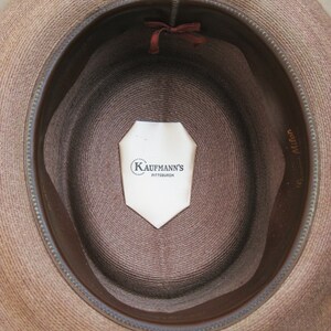 Pu&ograve; includere: L'interno di un cappello di paglia marrone con una fascia in pelle marrone. Un'etichetta bianca con il testo "Kaufmann's Pittsburgh" &egrave; attaccata all'interno del cappello.