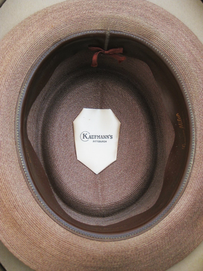Pu&ograve; includere: L'interno di un cappello di paglia marrone con una fascia in pelle marrone. Un'etichetta bianca con il testo "Kaufmann's Pittsburgh" &egrave; attaccata all'interno del cappello.