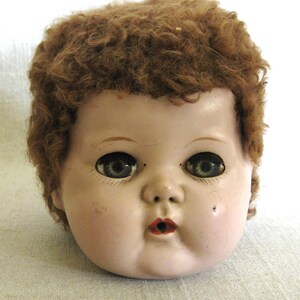 May include: Vintage doll head with a painted face, including blue eyes, red lips, and a beauty mark. The doll has brown, curly hair. The doll's face is a pale pink color.