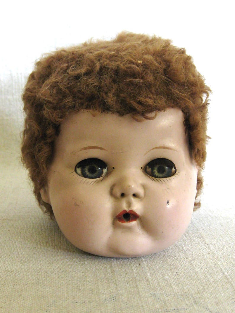 May include: Vintage doll head with a painted face, including blue eyes, red lips, and a beauty mark. The doll has brown, curly hair. The doll's face is a pale pink color.