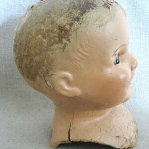 May include: Side profile of an antique doll head. The head is painted a pale flesh tone with a cracked, aged appearance. The doll has a small ear, a closed mouth, and a single visible green eye.