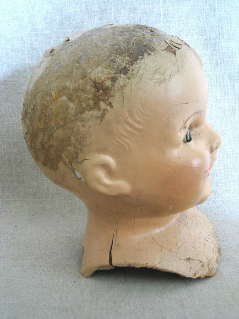 May include: Side profile of an antique doll head. The head is painted a pale flesh tone with a cracked, aged appearance. The doll has a small ear, a closed mouth, and a single visible green eye.