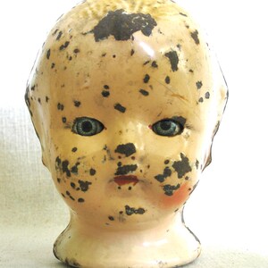 May include: Vintage doll head with blue eyes, red lips, and a pale complexion. The doll head has black spots and areas of paint loss, giving it a distressed appearance. The head is mounted on a short, rounded neck.