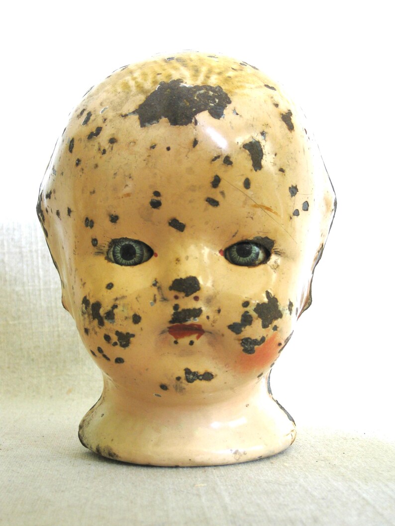 May include: Vintage doll head with blue eyes, red lips, and a pale complexion. The doll head has black spots and areas of paint loss, giving it a distressed appearance. The head is mounted on a short, rounded neck.