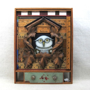 May include: A wall-mounted wooden shadow box featuring a central carved owl figure framed by dark brown wooden leaves. The background consists of vintage yellow measuring rulers with black text. A small green drawer with two knobs sits below.