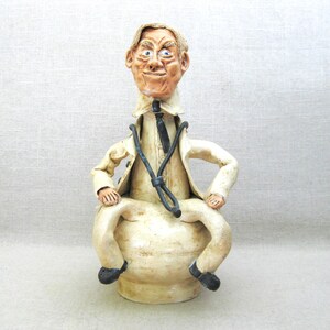May include: A ceramic figurine of a doctor wearing a white coat and a stethoscope. The doctor is sitting on a round base and has a comical expression on his face.