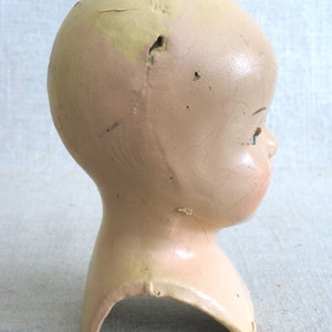 Puede incluir: Una cabeza de mu&ntilde;eca vintage hecha de yeso de color rosa p&aacute;lido y crema. La cabeza tiene una superficie agrietada y un agujero en la parte superior. La cabeza de la mu&ntilde;eca no tiene ojos y tiene la boca ligeramente abierta.