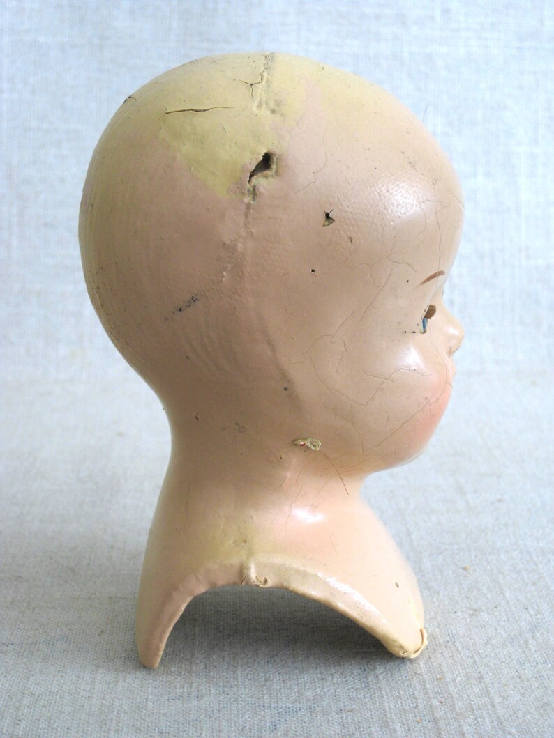 Puede incluir: Una cabeza de mu&ntilde;eca vintage hecha de yeso de color rosa p&aacute;lido y crema. La cabeza tiene una superficie agrietada y un agujero en la parte superior. La cabeza de la mu&ntilde;eca no tiene ojos y tiene la boca ligeramente abierta.