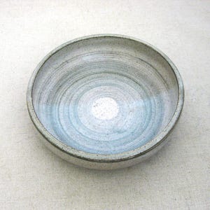 May include: A handmade ceramic bowl with a light blue and gray spiral design. The bowl has a circular shape and a textured surface. The interior features a white center. This decorative bowl is suitable for serving food or as a decorative piece.