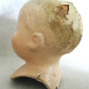 May include: A vintage doll head with a cracked, aged appearance. The head is a pale peach color with detailed facial features and remnants of painted hair. The top of the head has a large section missing, revealing the interior.