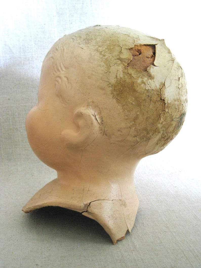 May include: A vintage doll head with a cracked, aged appearance. The head is a pale peach color with detailed facial features and remnants of painted hair. The top of the head has a large section missing, revealing the interior.