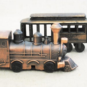 May include: Two antique-style metal train models. One is a locomotive, and the other is a cable car. The cable car has the text "Powell & Mason St." and "San Francisco" on it. Both models are bronze-colored.