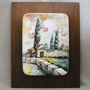 May include: A ceramic wall plaque with a painted scene of a small house with a brown roof and a green door, surrounded by trees and a stone wall. The sky is painted in shades of blue, pink, and yellow.