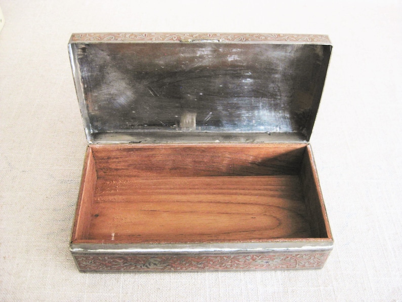 May include: An antique wooden box with a silver-colored lid. The lid is decorated with an intricate floral pattern. The box is open and the inside is lined with wood.