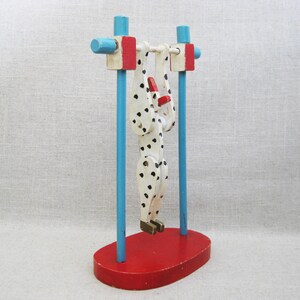 May include: A wooden toy figure of a person with black and white spots hanging from a blue and red wooden bar. The figure is painted white with black spots and has a red hat. The toy is on a red base.