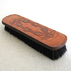 May include: A brown leather brush with a decorative design. The brush has black bristles and is made of leather.