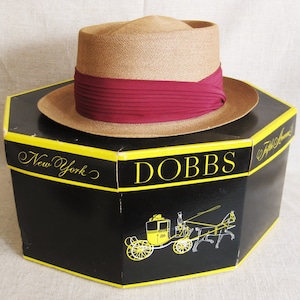Pu&ograve; includere: Un cappello di paglia marrone con un nastro color borgogna, seduto su una scatola nera con le parole "DOBBS" e "New York" stampate in giallo. La scatola presenta anche un'illustrazione in bianco e nero di una carrozza trainata da cavalli.