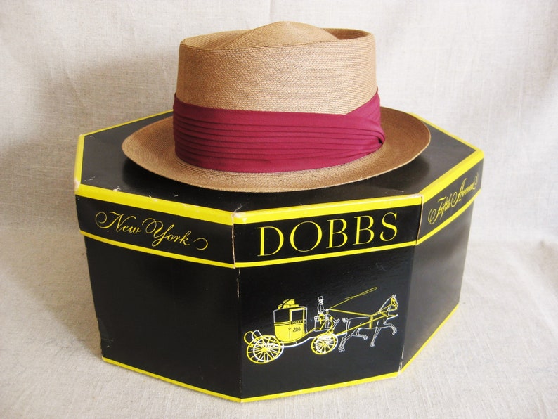 Pu&ograve; includere: Un cappello di paglia marrone con un nastro color borgogna, seduto su una scatola nera con le parole "DOBBS" e "New York" stampate in giallo. La scatola presenta anche un'illustrazione in bianco e nero di una carrozza trainata da cavalli.