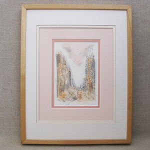 May include: A framed watercolor painting of a city scene with two tall buildings in the center. The buildings are rendered in shades of gray and brown, with a pink sky in the background. The painting is framed in a light wood frame.