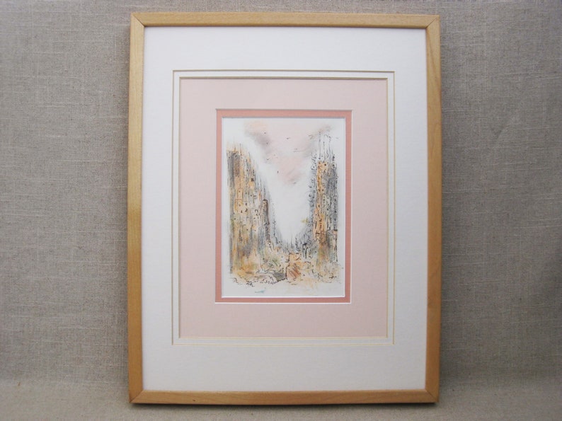 May include: A framed watercolor painting of a city scene with two tall buildings in the center. The buildings are rendered in shades of gray and brown, with a pink sky in the background. The painting is framed in a light wood frame.