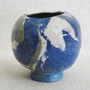 May include: A small, round ceramic vase with a blue, white, and gray marbled glaze. The vase has a wide opening and a small, round base.