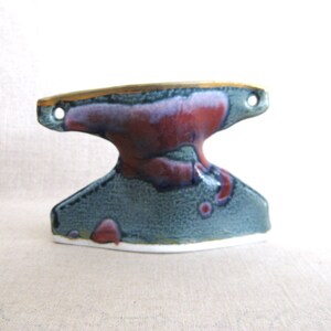 May include: A ceramic wall hook with a unique, abstract design. The hook is glazed in shades of blue, green, and red, with a gold accent on the top. It has two holes for hanging.