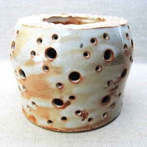 May include: A ceramic candle holder with a cylindrical shape. The off-white and brown glazed surface features numerous irregularly sized holes. The top edge has a scalloped design. The pottery has a rustic, handmade appearance.