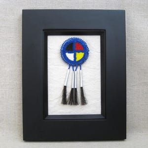 May include: A framed beaded medallion with a blue, black, red, and yellow design. The medallion has three black tassels hanging below. The frame is black and the background is white.