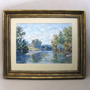 May include: A framed landscape painting featuring a serene river scene. The artwork depicts a house on a distant shore, surrounded by trees with colorful foliage. The painting is set within a gold-colored ornate frame.