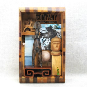 May include: A wooden shadow box with a variety of carved wooden figures and abstract elements. The box features a checkerboard pattern on the frame and includes the word "COMPANY" in the upper section. The box contains a small wooden deer, a carved figure, and a wooden head.